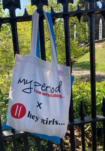 My Period X Hey Girls branded cotton tote bag hanging on the railings in a park in Edinburgh