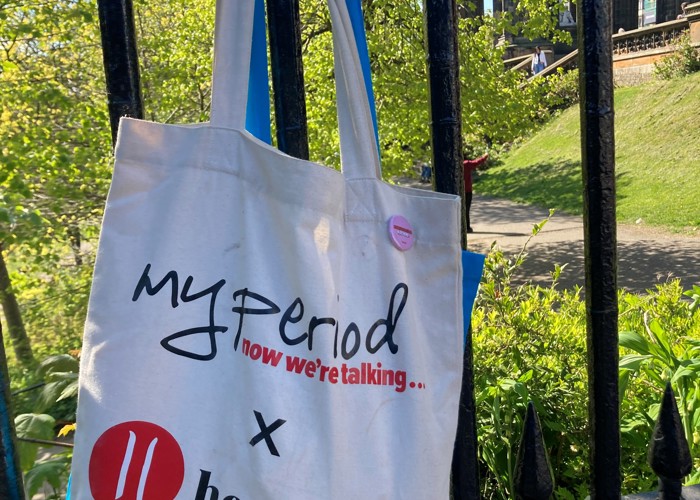 My Period X Hey Girls branded cotton tote bag hanging on the railings in a park in Edinburgh