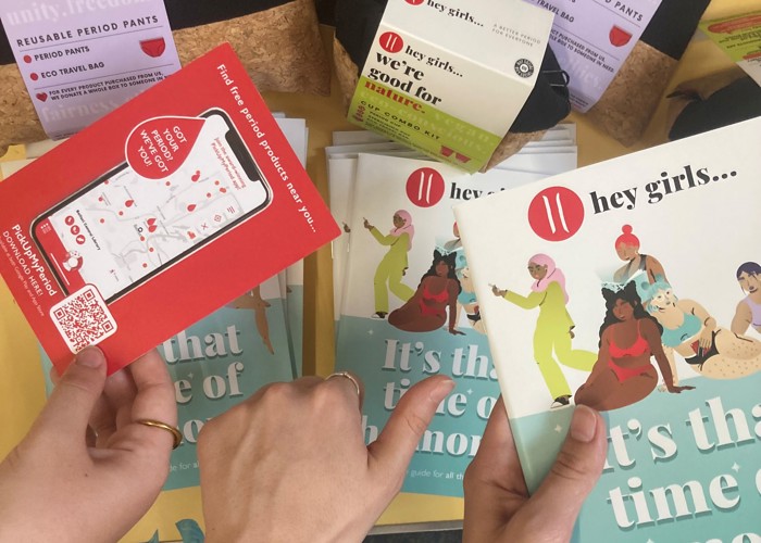 Three hands picking up the Its that time of the month booklet, PickUpMyPeriod app postcard and a thumbs up with the background of the My Period conversation table resources and period products