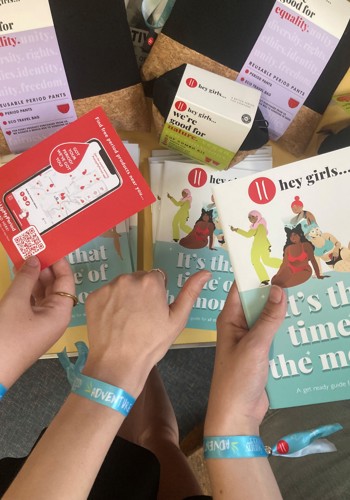 Three hands picking up the Its that time of the month booklet, PickUpMyPeriod app postcard and a thumbs up with the background of the My Period conversation table resources and period products